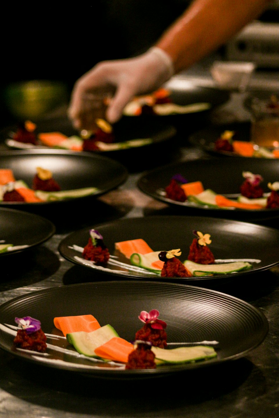 Mano enguantada de chef arreglando aperitivos gourmet en platos oscuros.